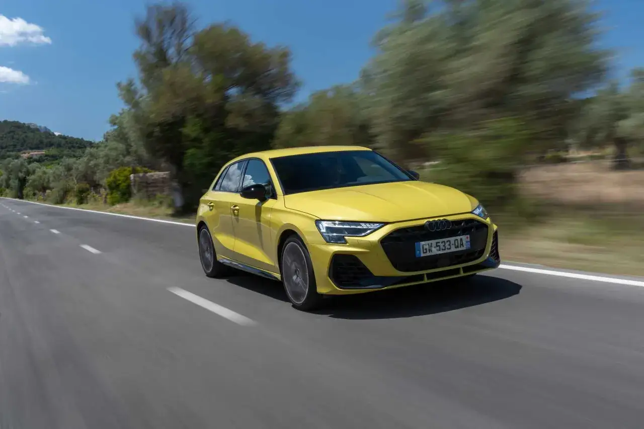 Une Audi S3 jaune vif, pleine de chevaux, file sur une route sinueuse bordée d'arbres.