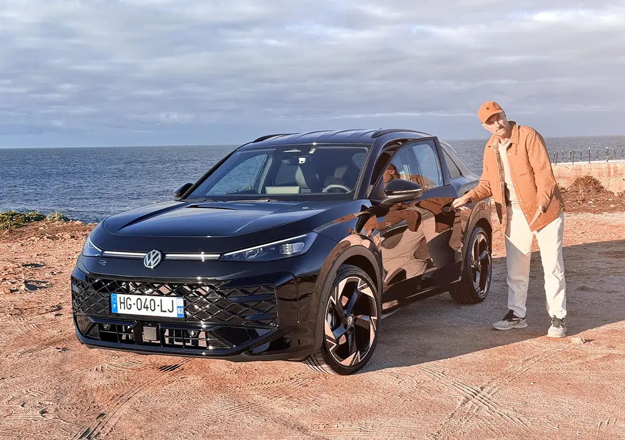 Un homme pose près d'un Volkswagen T-Roc noir, prêt à découvrir son moteur. La mer et le ciel bleu en arrière-plan.