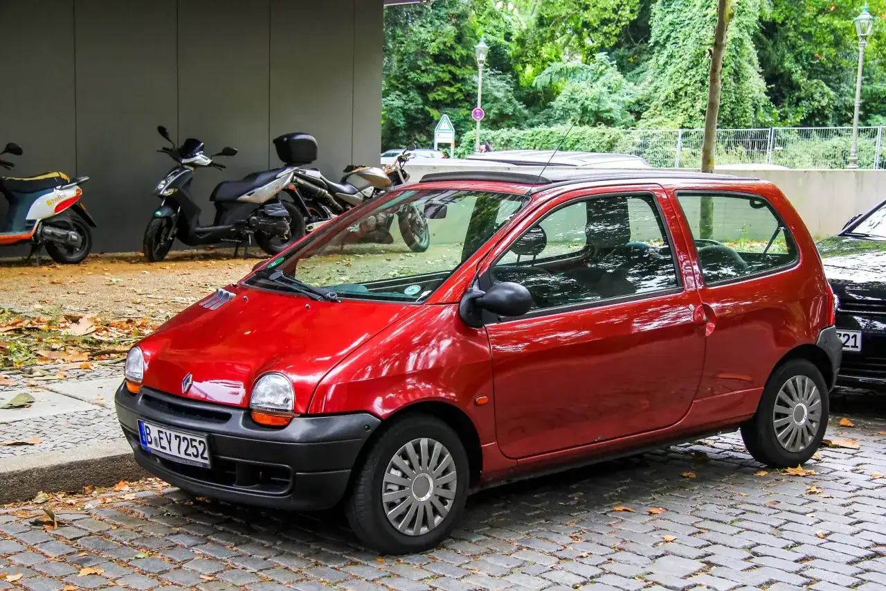 Twingo 1 rouge gar&eacute;e sur pav&eacute;s, avec des scooters et des arbres en arri&egrave;re-plan. Pensez &agrave; v&eacute;rifier le filtre habitacle Twingo 1 pour un air pur.