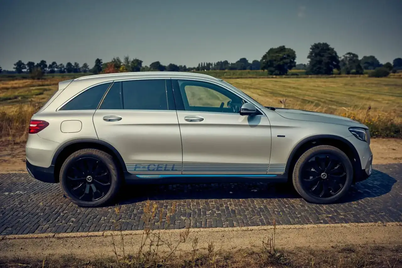 Voiture argentée, un dimension GLC coupé avec des bandes bleues sur le flanc, garée sur une route pavée devant un paysage champêtre.
