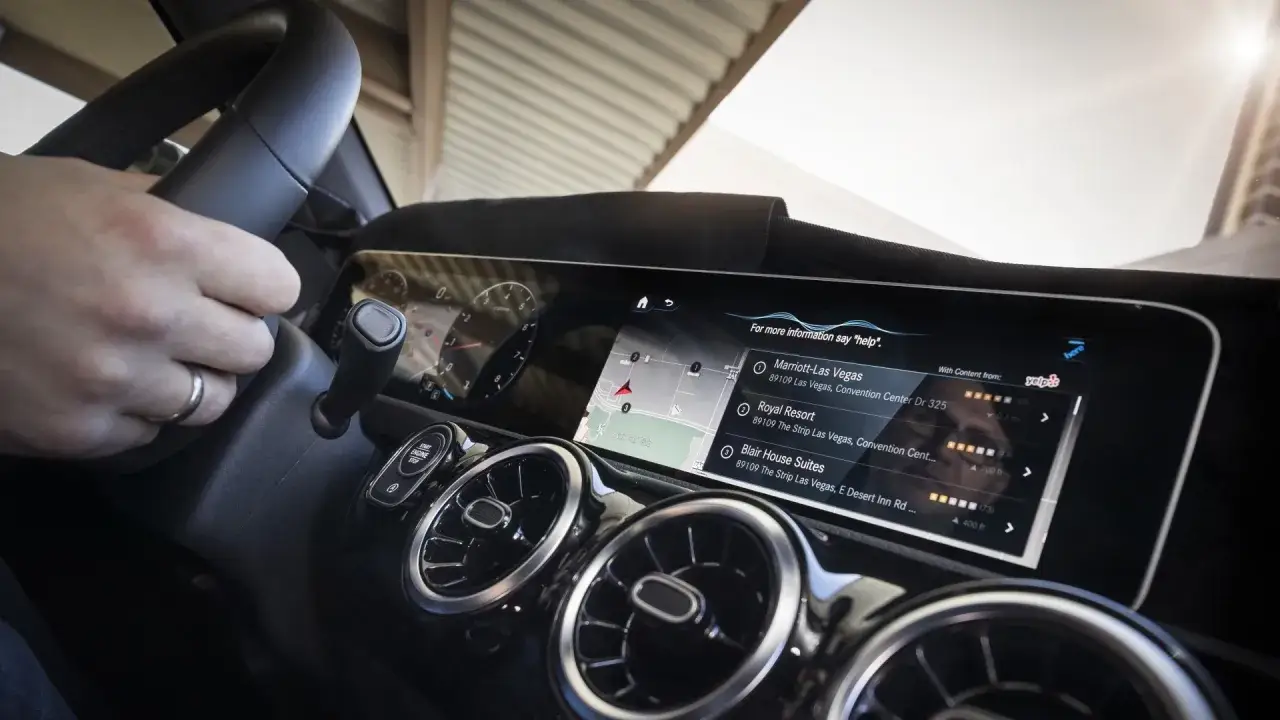 Vue de l'intérieur d'une Mercedes Classe A, avec une main sur le volant et l'écran tactile affichant une carte et des suggestions d'hôtels.