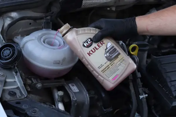 Main d'une personne portant un gant noir verse du liquide de refroidissement rose dans le réservoir d'un moteur de voiture.