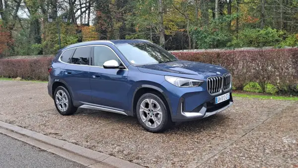 Avis sur la BMW X1 sDrive 18d 150 ch : ce SUV bleu est garé sur un chemin pavé, entouré d'arbres aux couleurs d'automne.