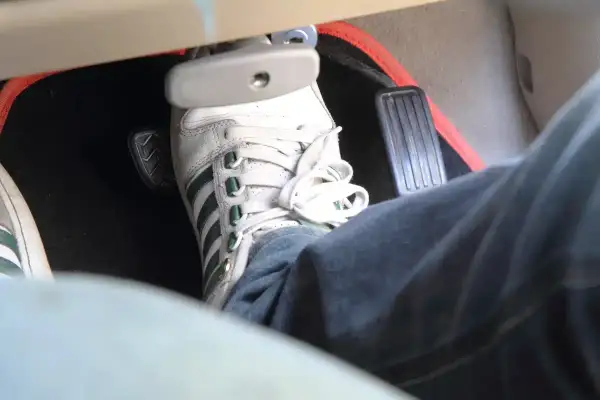 Un pied en basket Adidas appuie sur la pédale de frein molle. Le tapis de sol rouge est visible.