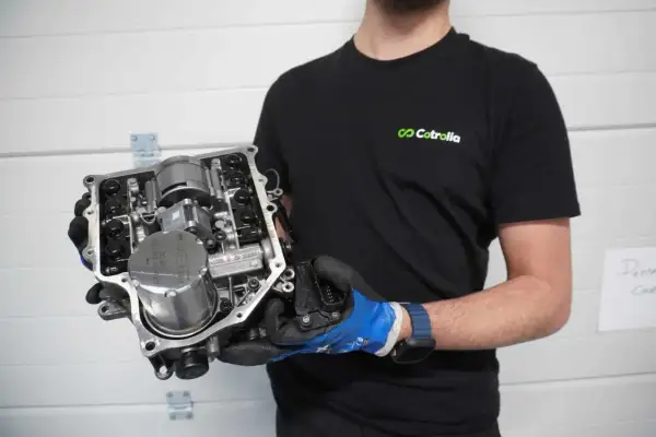 Un homme en t-shirt noir tient un ensemble de transmission complexe, montrant le levier de vitesse et d'autres composants mécaniques.