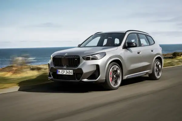 Un essai BMW X1 gris argenté roule sur une route côtière, l'océan bleu en arrière-plan.