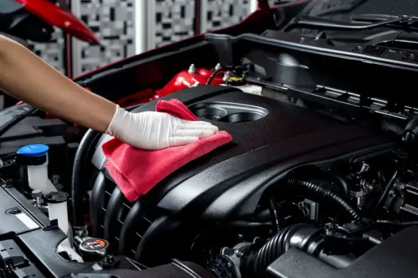 Main d'une personne portant un gant blanc, essuyant le capot du moteur d'une voiture avec un chiffon rouge pour nettoyer l'huile moteur.